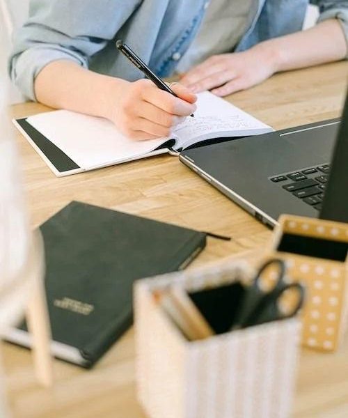 A copywriter sitting in front of a laptop, writing in a notebook